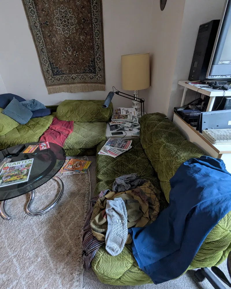 Sitzecke vorher: Kleidung, Decken und Zeitschriften auf der grünen Sitzgarnitur.