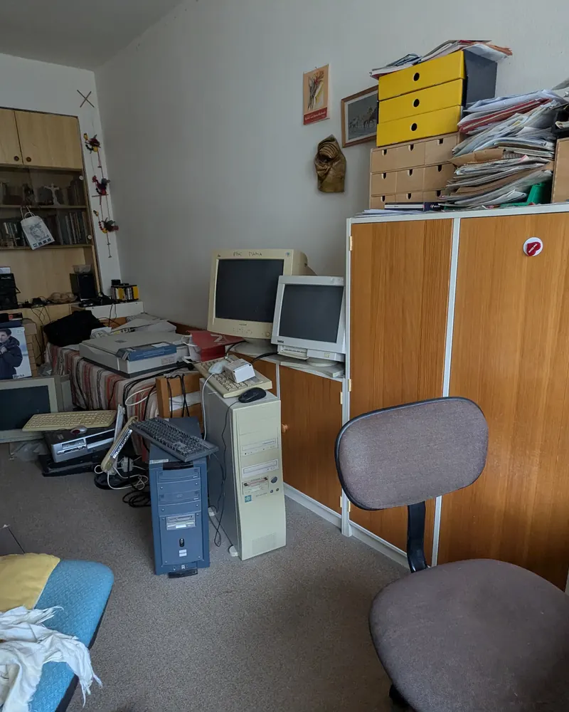Arbeitszimmer vor dem Aufräumen: alte Computer, Tastaturen und Papierstapel auf Kästen und Boden.